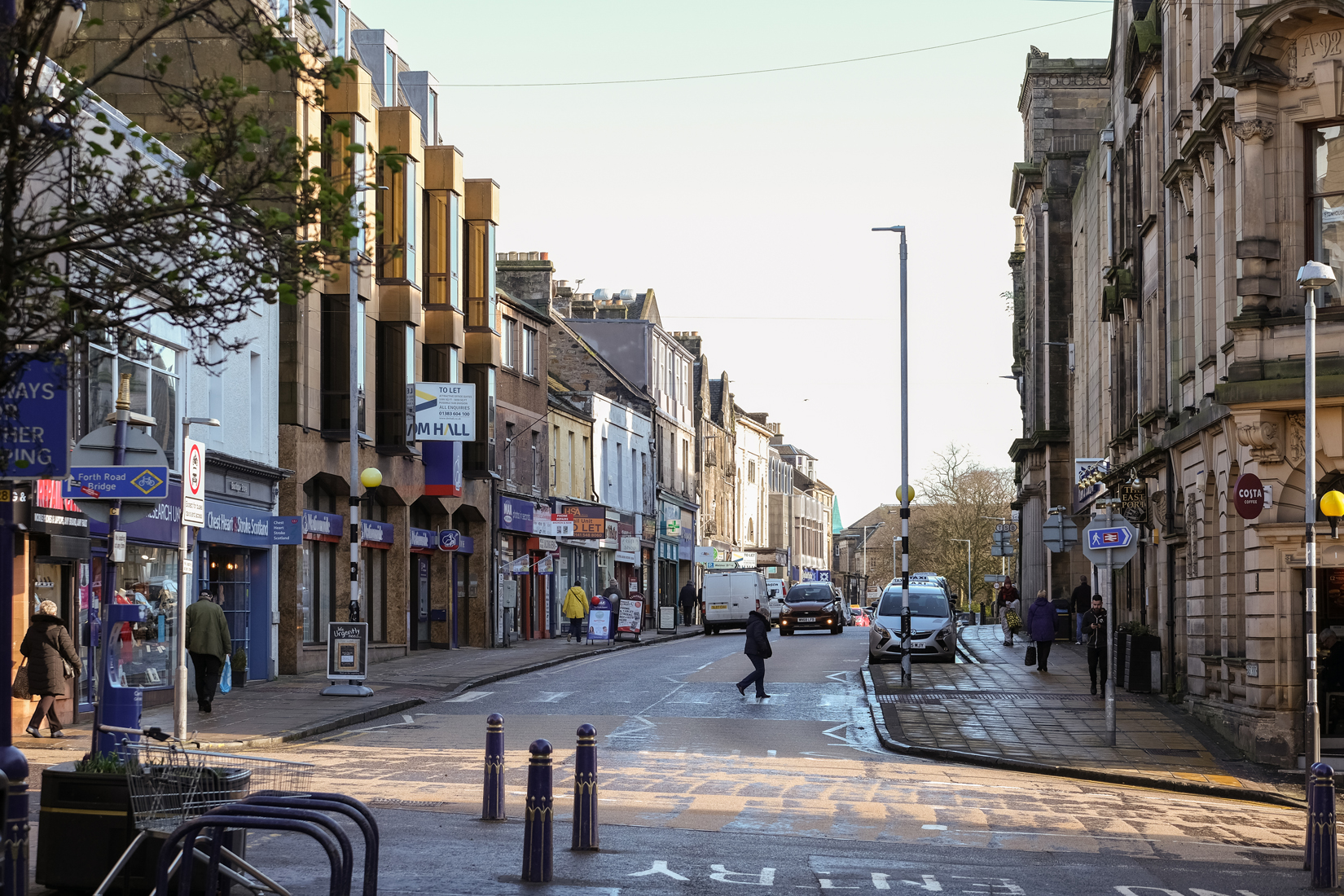 High street with pavements, car driving towards and pedestrian on zebra crossing. Buildings on right look over 100 years old of stone and the left are more modern.