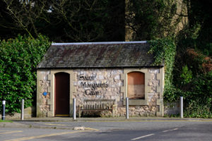 Small building surrounded by tress and road in front. It has writing Saint Margaret's Cave with doorway on left and window on right