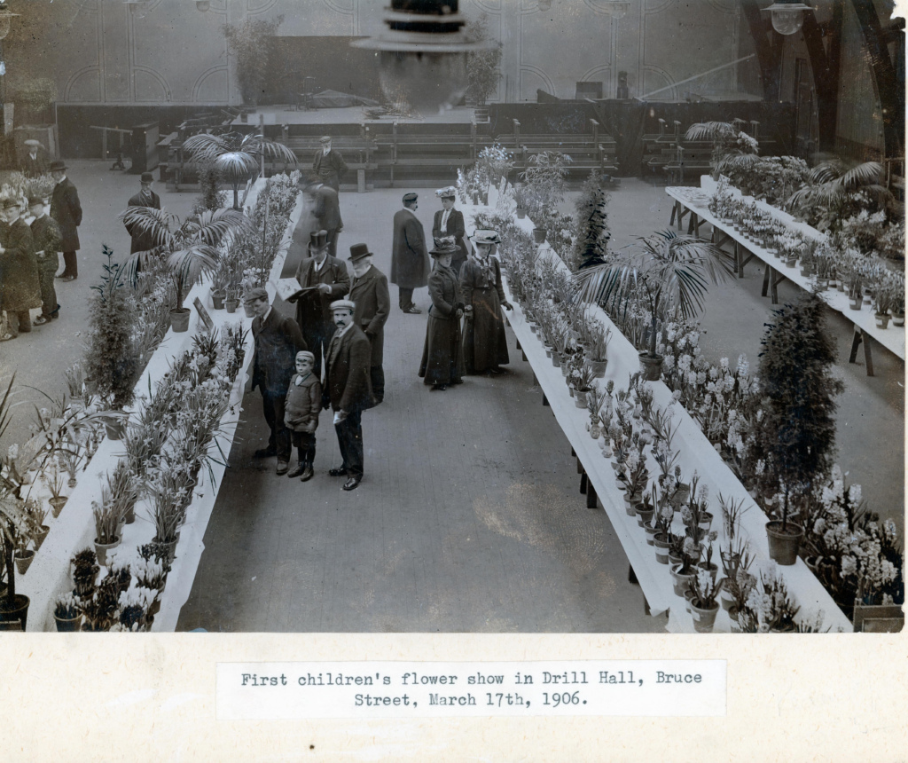 Black and white image dated 1906 in spacious hall. Group of 10 people in centre, well dressed mixture ladies, gentlement and a child with some looking at the camera. Three long tables from bottom of the picture up are covered in various plants and flowers. Caption on the image 'First children's flower show in Drill Hall, Bruce Street, March 17th, 1906.'