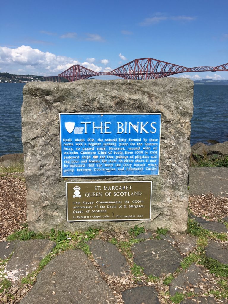 Stone with signs telling story of The Binks natural jetty and St Margaret Queen of Scotland. Behind is the sea and Forth Bridge.